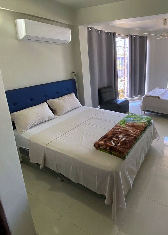 Doble suite: cama con respaldo azul, aire acondicionado y otra cama al fondo, junto al ventanal