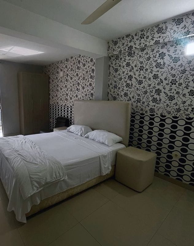 Suite individual: cama queen con respaldo tapizado, papel tapiz floral en blanco y negro, un puf y el ropero