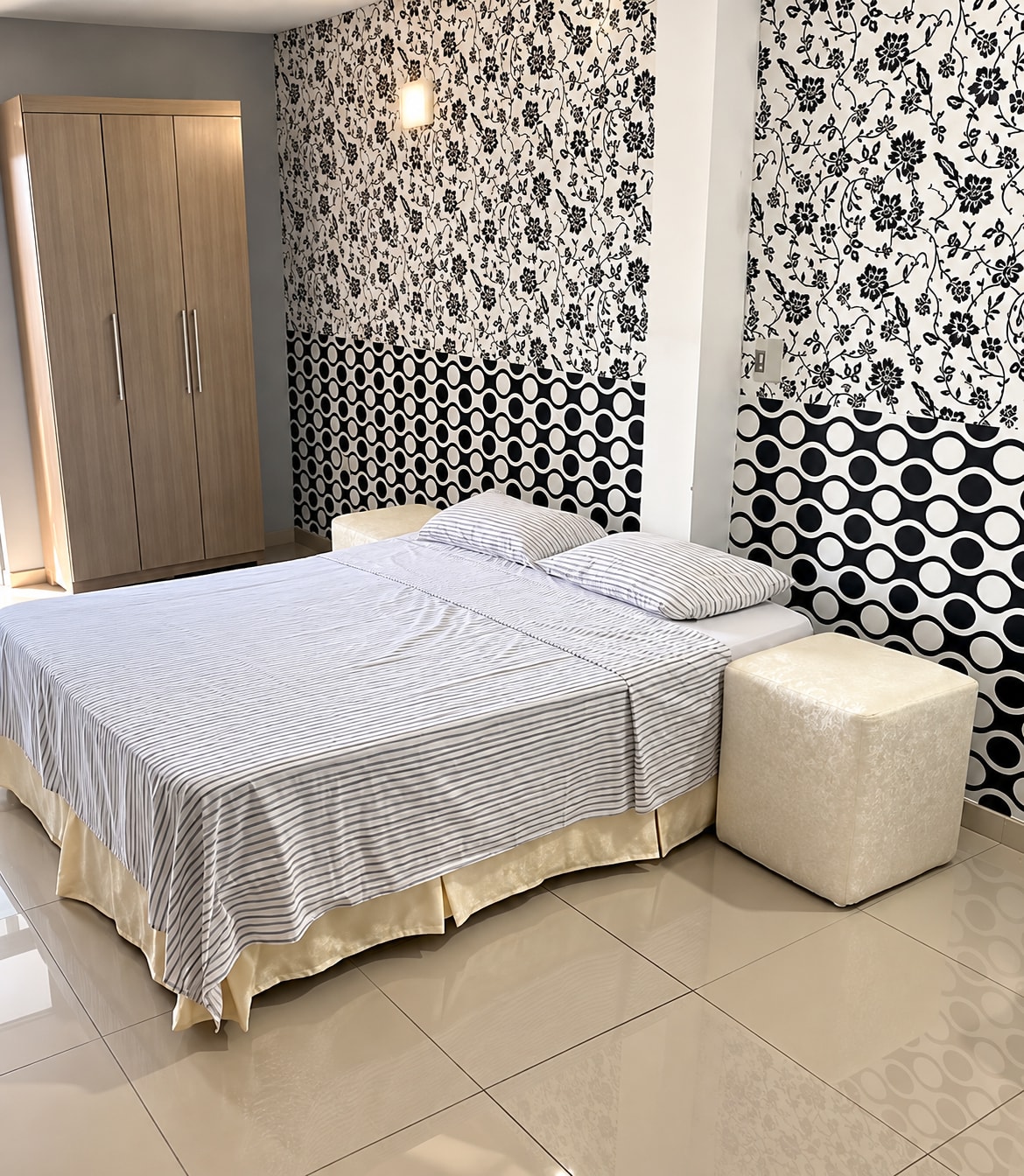Una suite del hotel con cama queen: la cama tendida, el papel tapiz floral en blanco y negro, el ropero y un puf